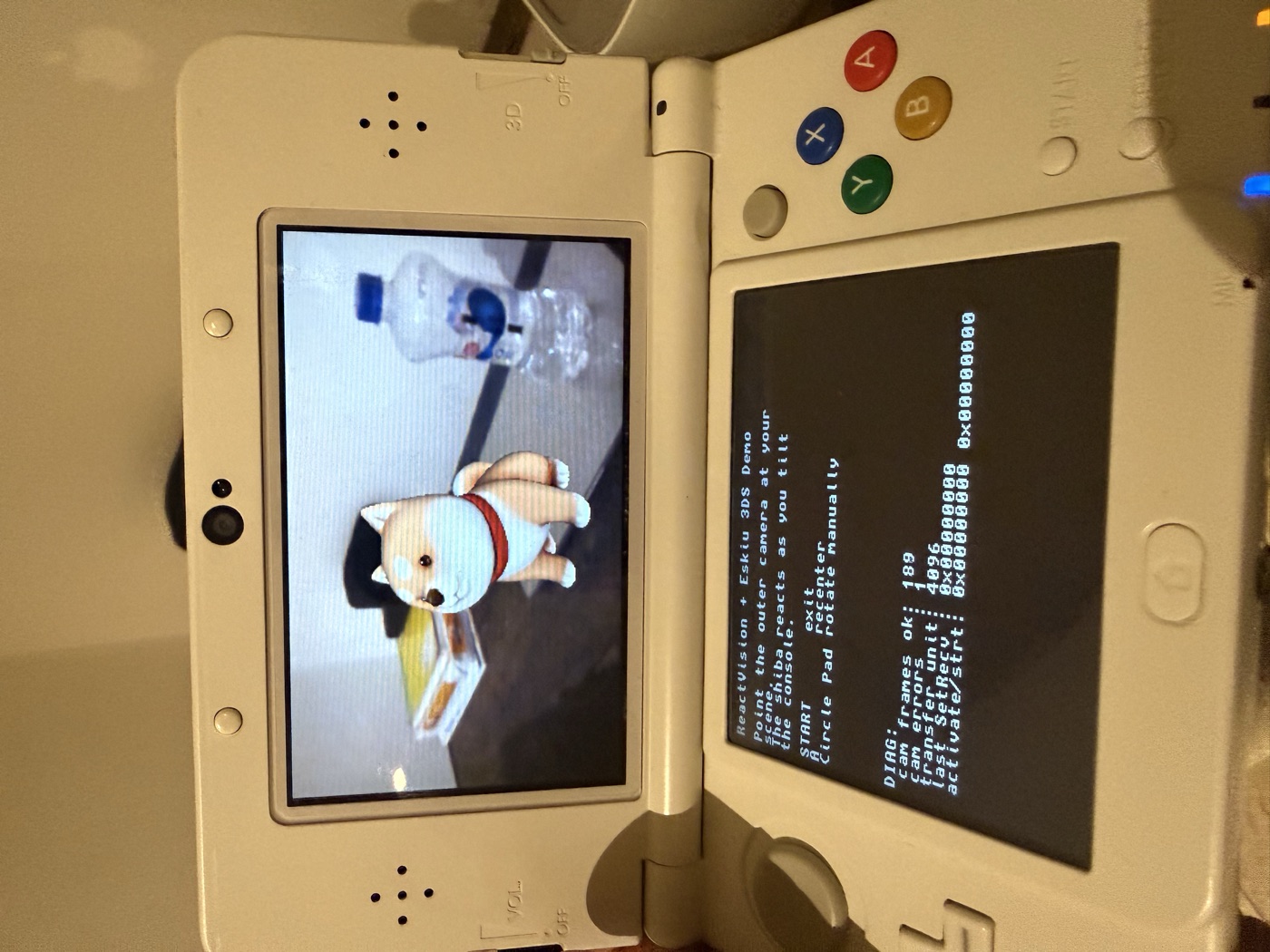 A New Nintendo 3DS running the ReactVision and Eskiu demo: a low-poly shiba 3D model composited over the live outer-camera feed on the top screen, with the demo's controls and per-frame diagnostics on the bottom screen.
